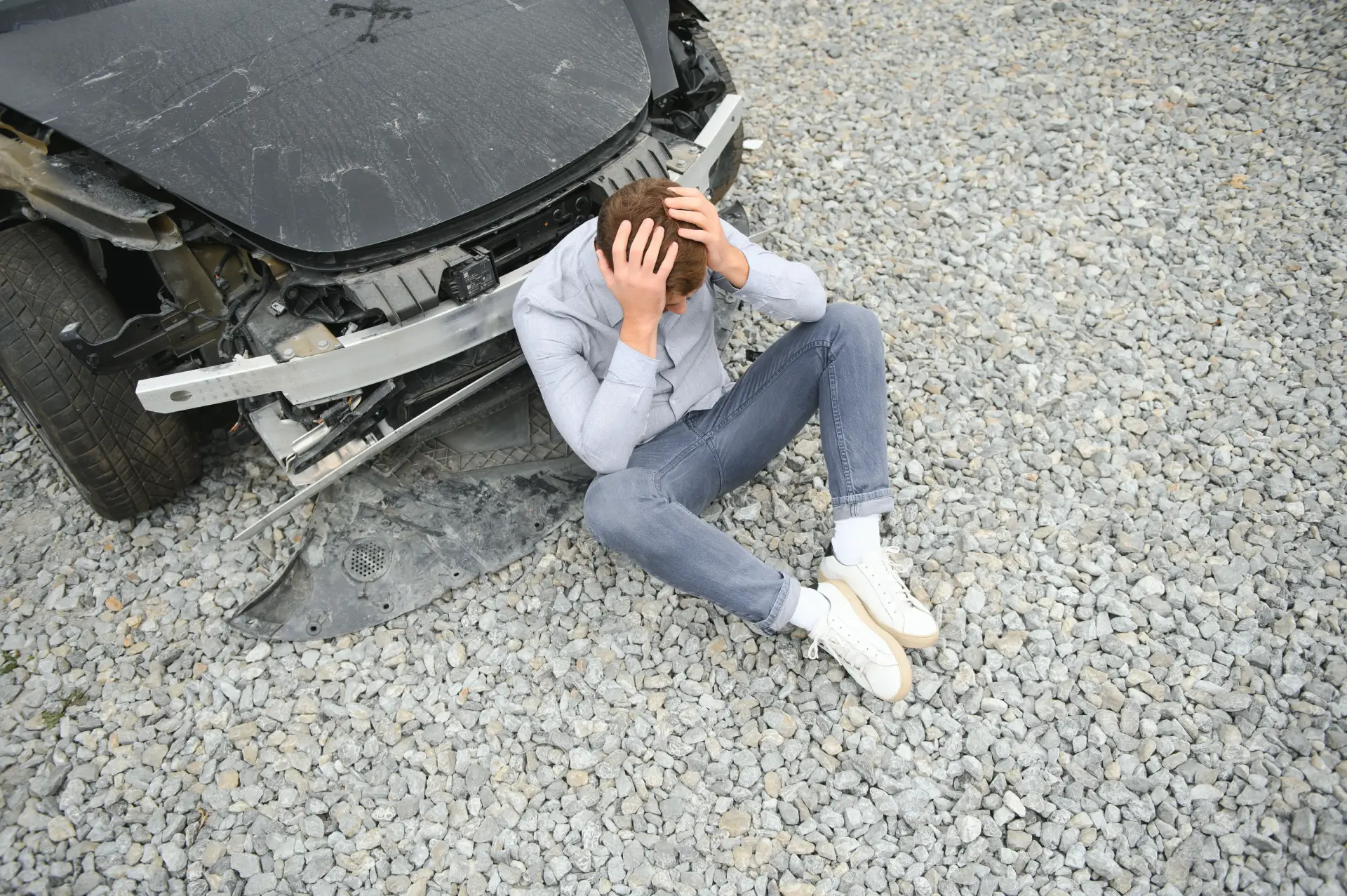 man holding his head sitting against a damaged car